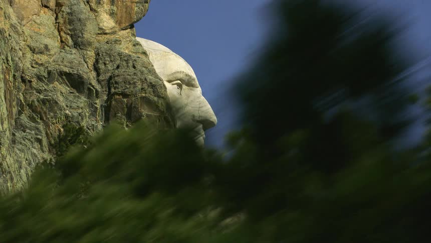Mount rushmore monument sits on mountain side near conifer trees under clear sky