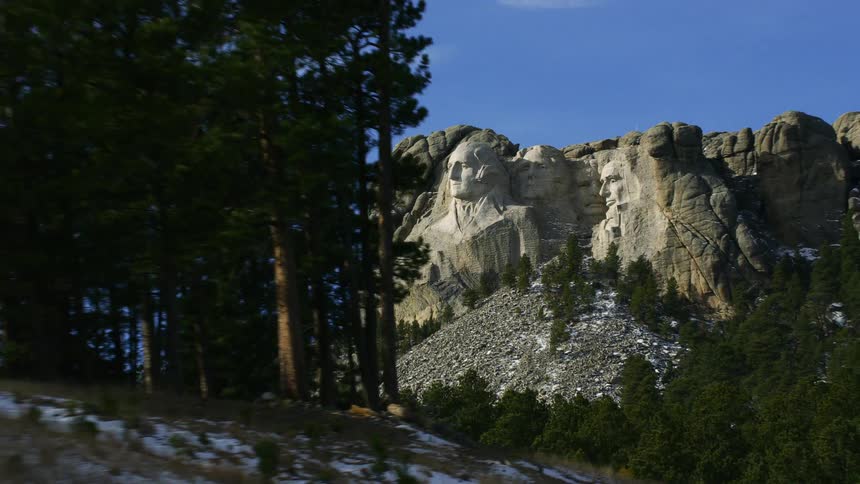 Clouds and contrails sit in sky over mount rushmore and forest occupying slope across road from monument