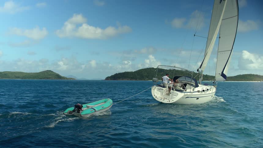 Small sailboat yatch towing an inflatable raft in front of islands