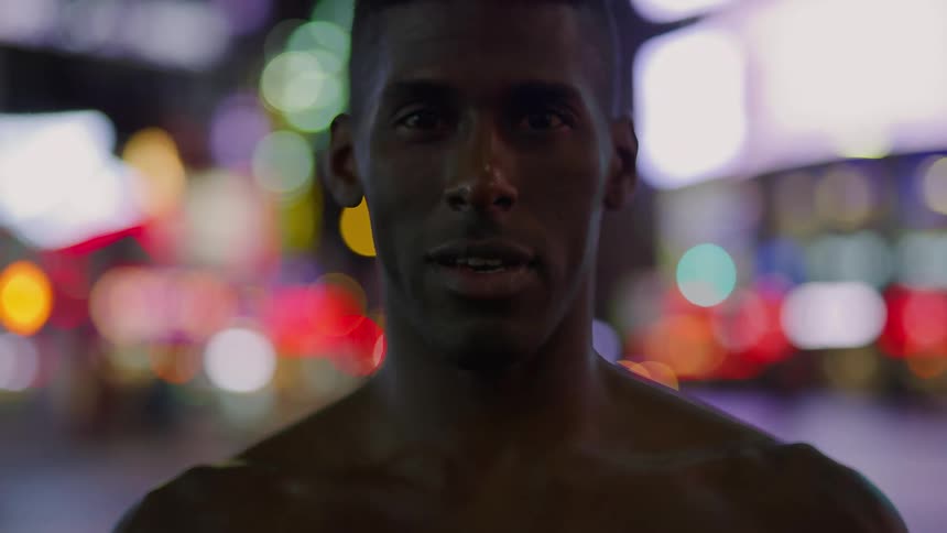 A black man takes a deep breath and looks forward and there are bright colored lights behind him