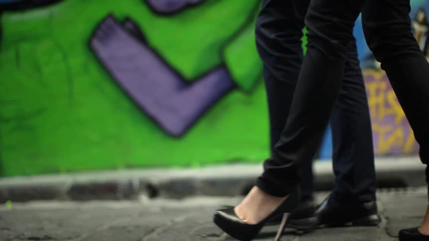 Leg view only of two people walking down a road past graffiti buildings