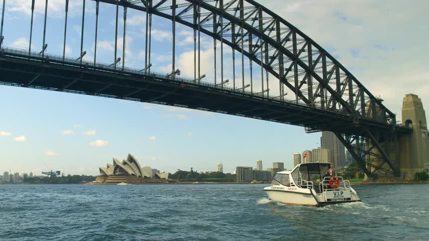 Boat makes a pass under sydney harbour bridge with a view of sydney opera house in the background