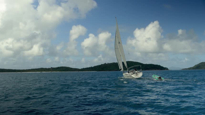 A sailboat is pulling a smaller boat in rough water under a cloudy sky