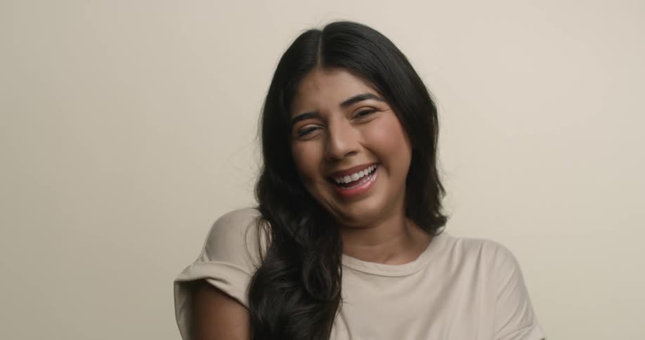 A young, ethnic female with long dark hair talks and throws her head back as she laughs while she looks at the camera with a big smile