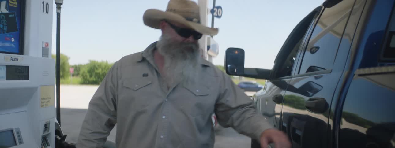 Bearded man wearing sunglasses and a hat stops at a gas station to fill the tank of a black truck