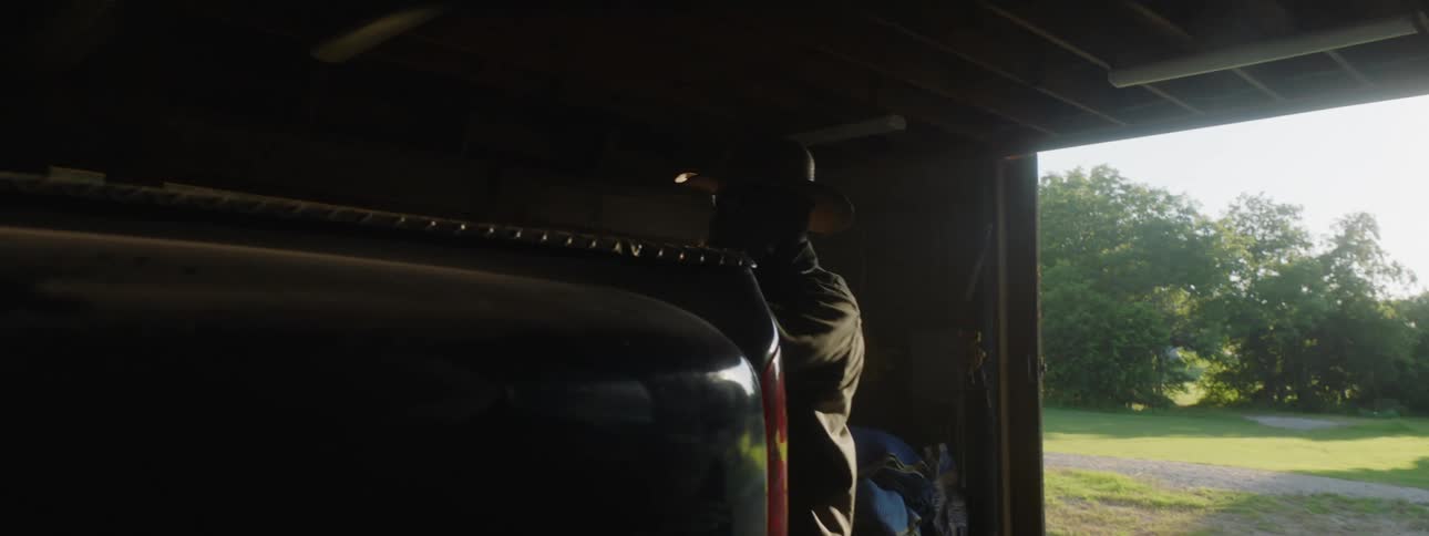 Bearded man wearing a hat opens a barn door and starts loading a truck at morning