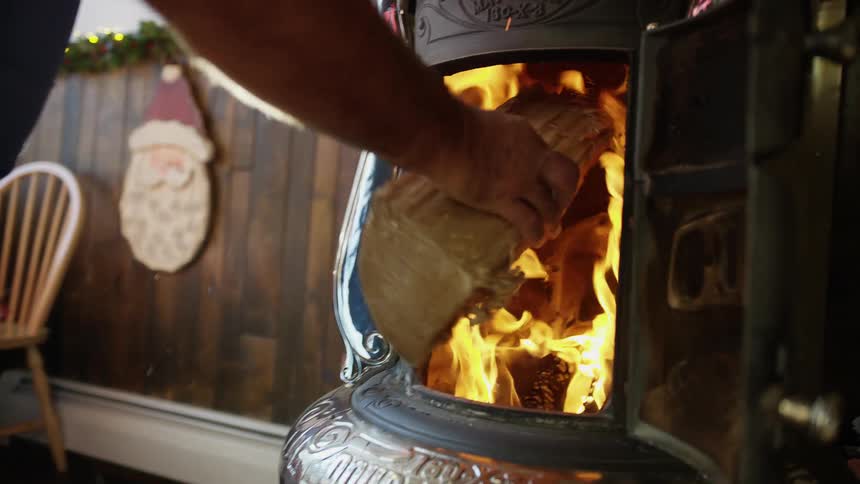 A man in his home is tossing a log into an active wood burning stove during the daytime in the christmas holiday season
