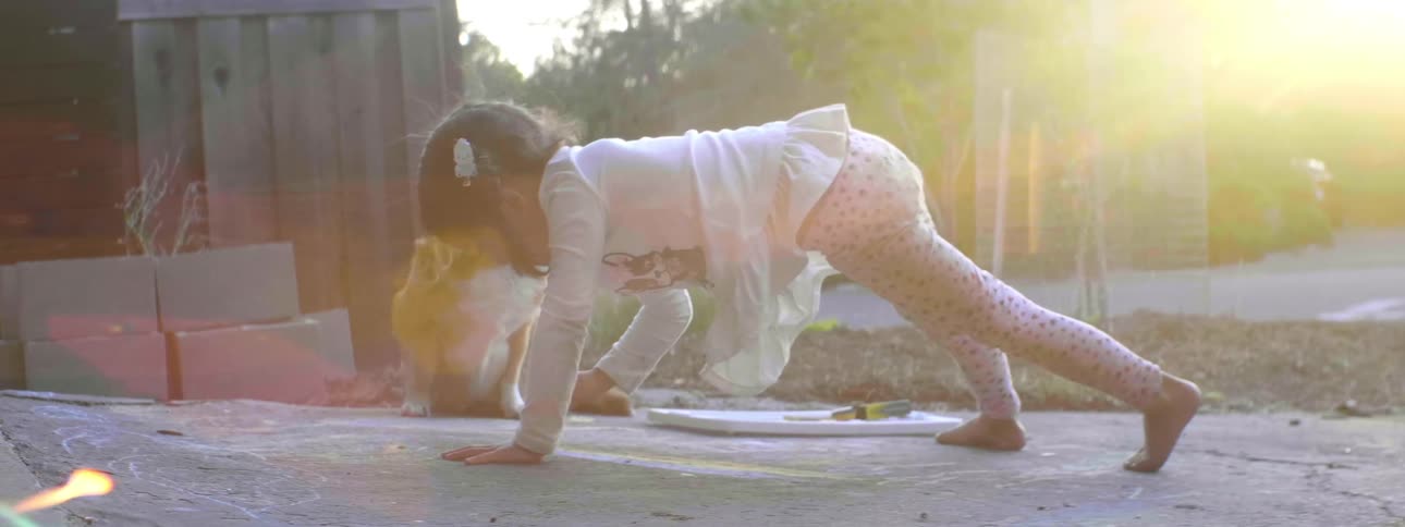 A toddler draws with chalk in her driveway while her dog waits nearby