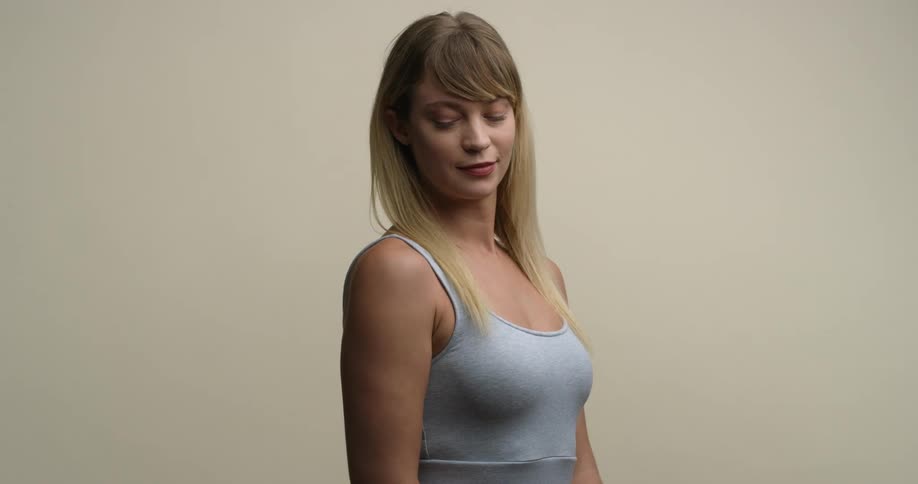 A young, blonde female model with grey tank top tucks her chin, looks down, and then back up at the camera with a slight smile.