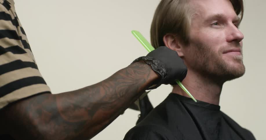 A tattooed black man wearing gloves uses scissors and a comb to cut a white, bearded blonde man's hair.