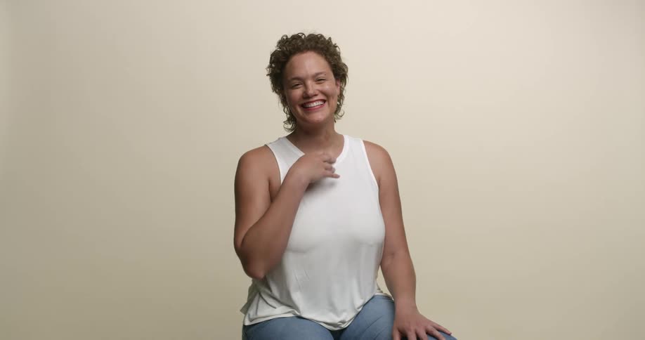 A young, curvy woman with short curly hair sitting for a portrait puts her hands to her chest and laughs