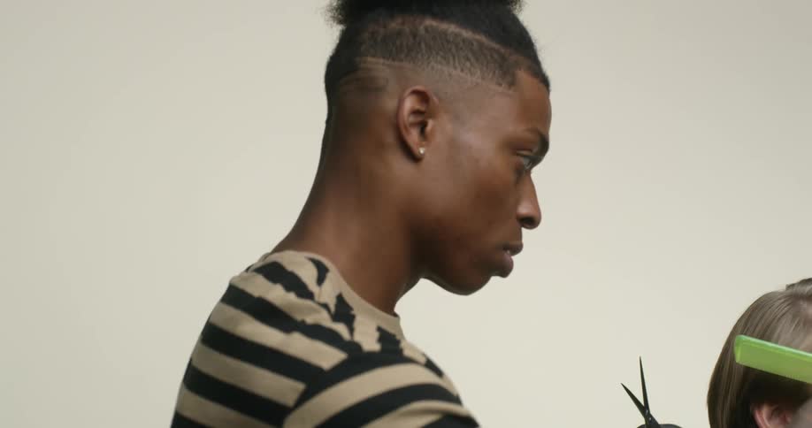 A young man of color is trimming and grooming a customer's hair in a barber chair.