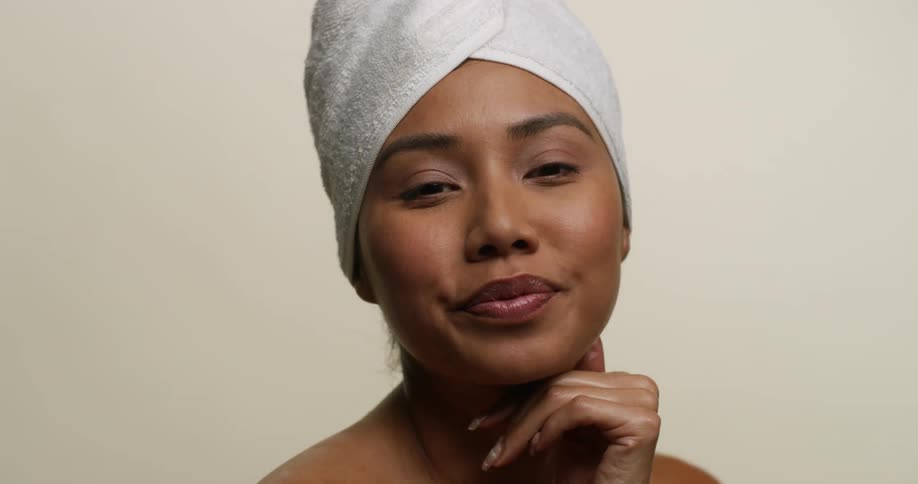 A young, ethnic woman wearing only a towel looks at the camera and smiles with pursed lips while caressing her neck with her hand.