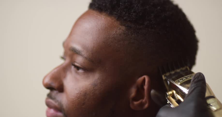 A young, black man with mustache is relaxed as he sits getting his hair trimmed by a person with an electric razor.
