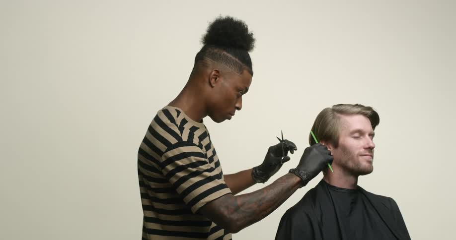 A blonde, white man is sitting in a barber chair with cape on receiving a haircut from a young left-handed black man with scissors and comb.