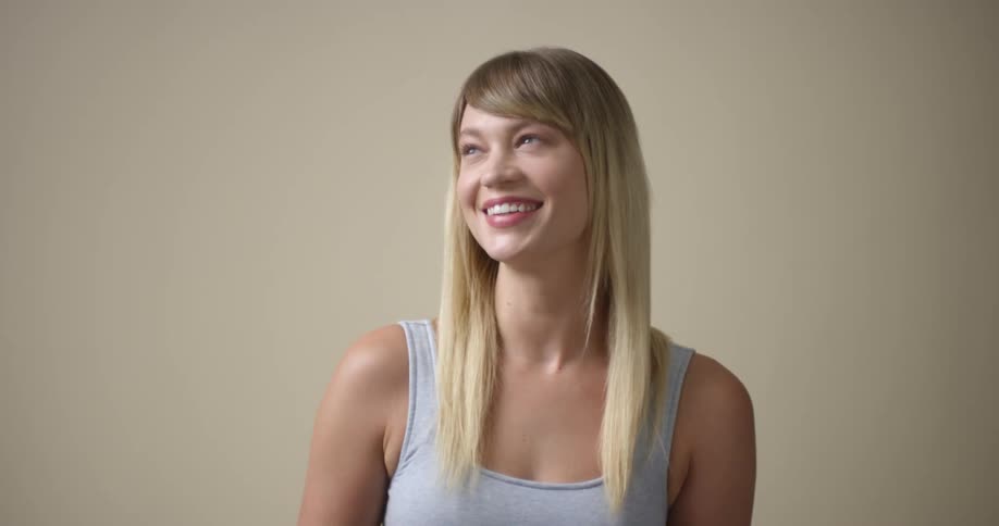 A young, blonde Caucasian woman with light eyes looks at the camera with a slight smile and then leans her shoulders forward and body to the side as she giggles, smiles, and looks away.
