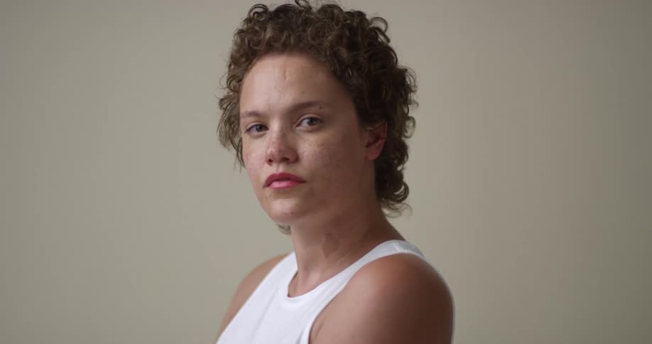 A young, white woman with short curly hair and freckles looks over her shoulder and then at the camera with a straight face.