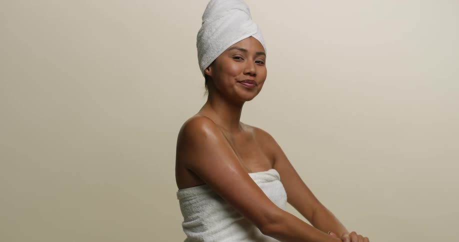 A young, Asian woman wearing only a towel is applying moisturizer to her skin after showering.