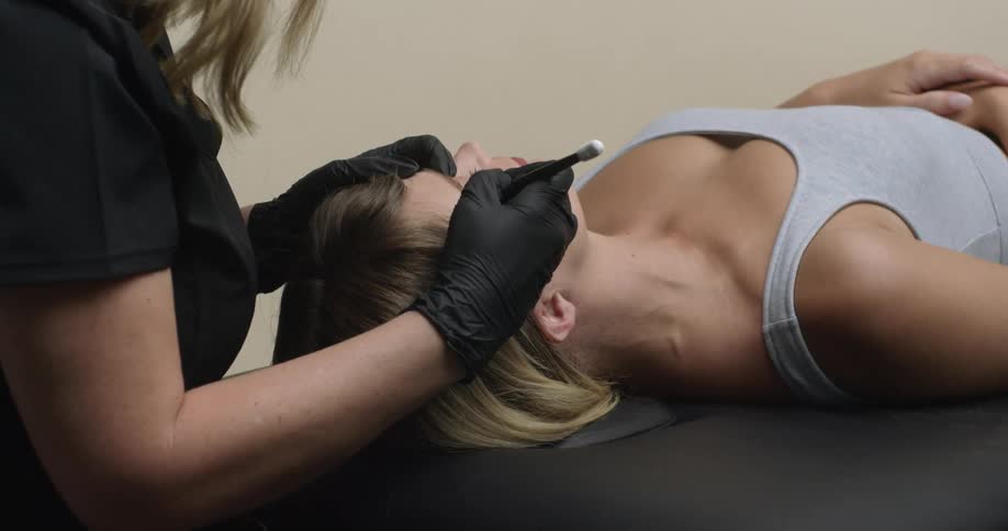 A young, blonde, white woman is lying on a spa table getting her eyebrows microbladed by a skilled esthetician.