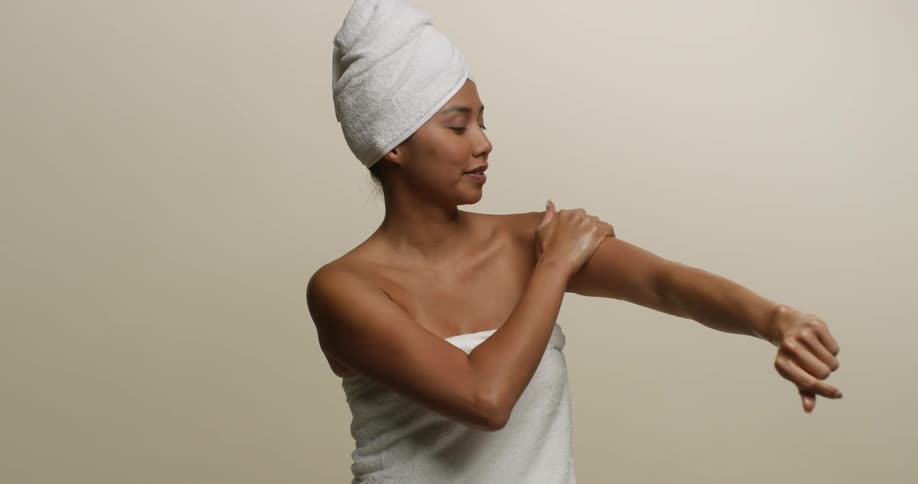 A young, Asian woman wearing a towel rubs lotion on her arms after showering.
