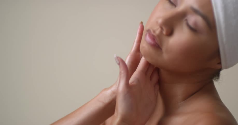 An Asian woman wearing a towel rubs her hands together and applies cream to her neck and smiles