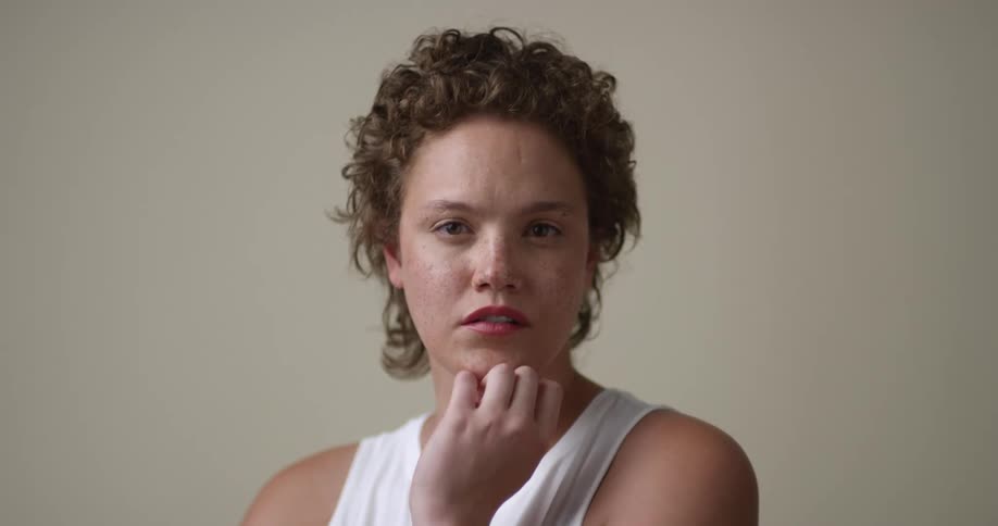 A young, freckled woman with short curly hair with her hand up to her chin turns to look at the camera.