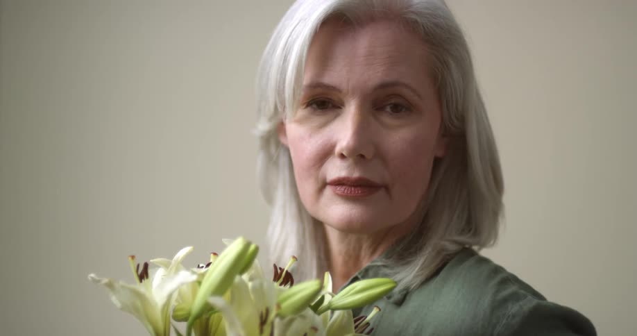 A mature white-haired Caucasian woman holding a bouquet of lily flowers opens her eyes and turns to look at the camera.