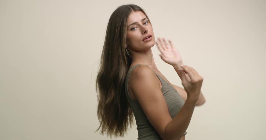 A young woman with light eyes and wearing a tank top raises her arms and runs her hands through her hair, tossing it back and through her fingers as she looks coyly at the camera.