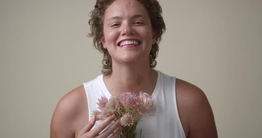 A young, white woman with short, curly hair and freckles gives a beaming smile to the camera while holding a bouquet of flowers.