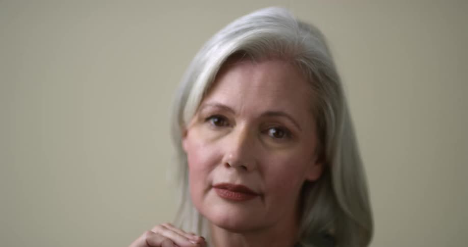 A mature Caucasian woman with white hair, dark brown eyes, and deep red lipstick leans on her hands looking away and then turns to look straight at the camera.