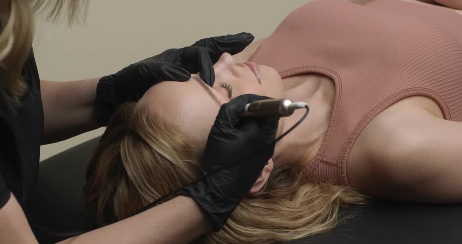 An eyebrow technician wearing black gloves is microblading the eyebrows of a young, blonde female customer who is laying on the spa table.