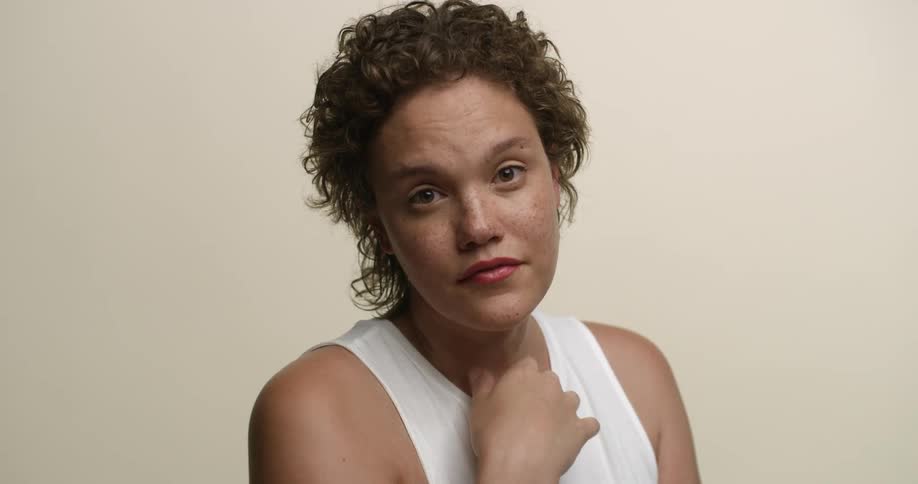 Close up of a young, freckled white female with boy-cut curly hair looking direct at the camera with her hand to her chin