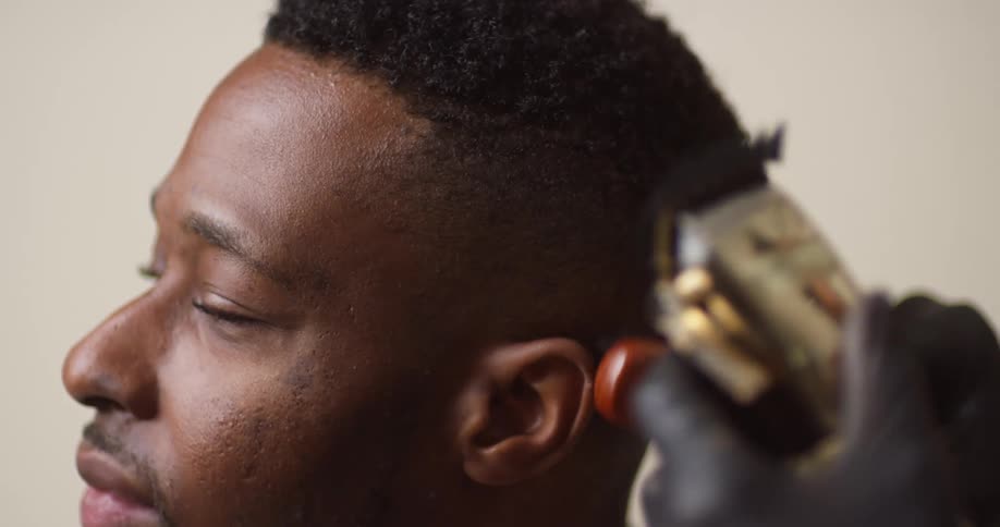 A young, black man in profile is having his hair trimmed by a person with an electric razor.