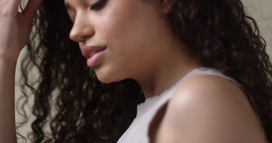 A young, ethnic female model pulls her hand through her dark, thick, curly hair as the camera pans down from profile to upper body.