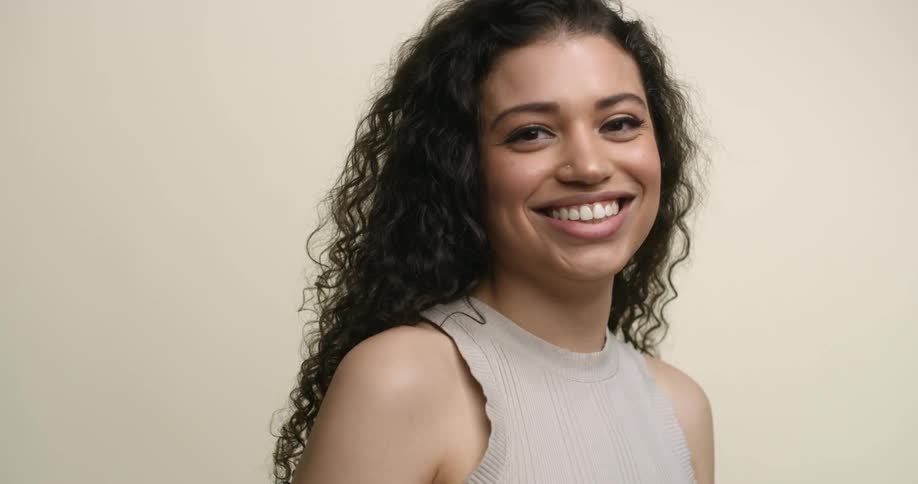 A young, multicultural woman with dark, curly hair, dark eyes, and a big beaming smiles poses for the camera in studio