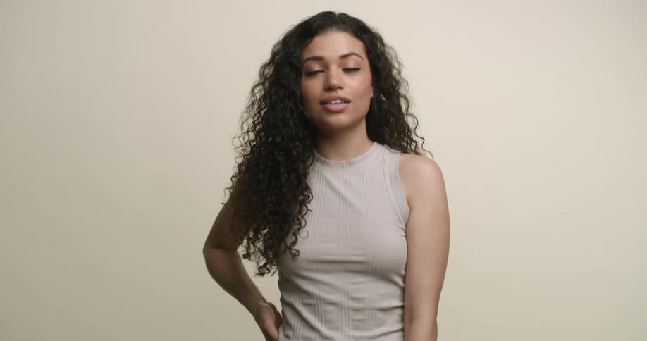 A young, multicultural woman with long, dark, curly hair moves her hand down her arm while posing for the camera and gives an alluring smile to the camera