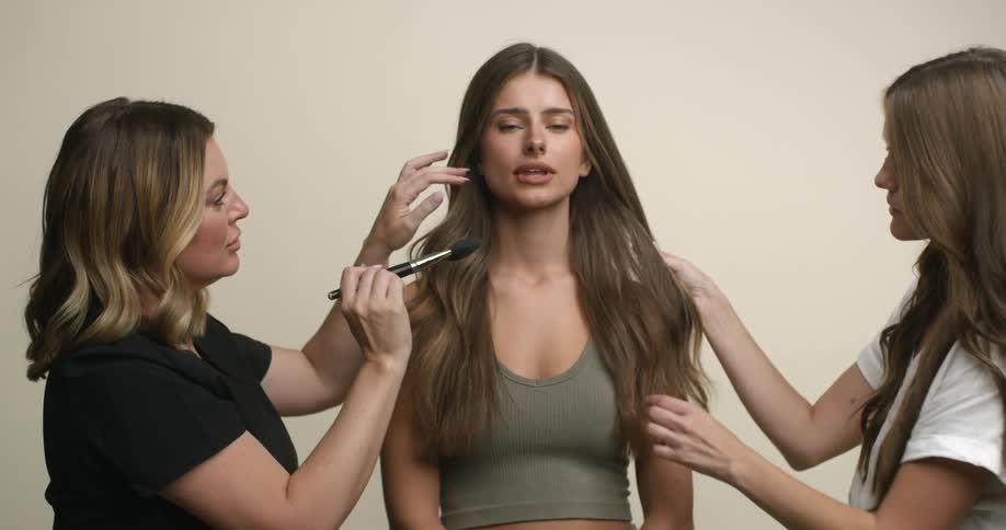 A young, Caucasian female model cooperatively stands still with an aloof expression as she is made up by a makeup artist and hair stylist.