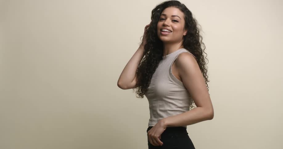 A young, multicultural woman with long, dark, curly hair tosses it back and up as she smiles and adjusts poses for the camera.