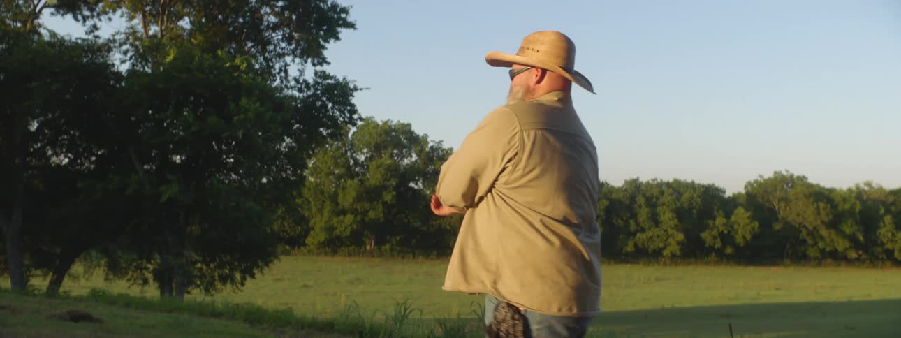 A heavy set man with a long beard and wearing a cowboy hat and sunglasses is rolling up his sleeves as he walks outside
