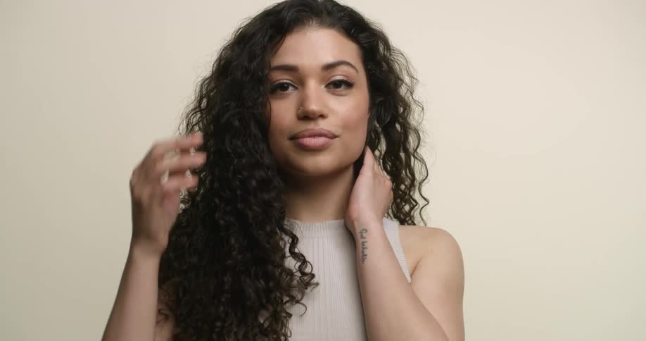 A young, female Pacific-Islander brushes her long, curly hair back with her hands and tosses it forward while giving a beaming smile to the camera