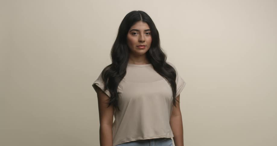 A young, ethnic woman with long, dark hair stands comfortably and gazes directly into the camera as it moves closer.