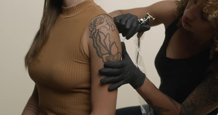 An ethnic female tattoo artist with brown curly hair is tattooing a rose on a caucasian woman's shoulder.