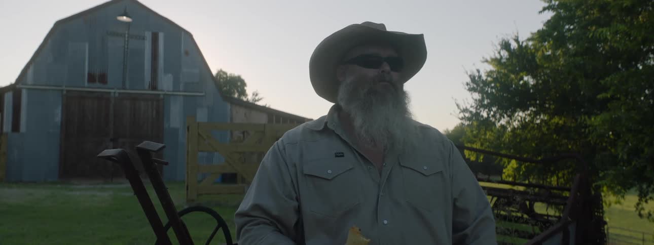 Man with cowboy hat sunglasses and beard eats sandwich looking around in the countryside