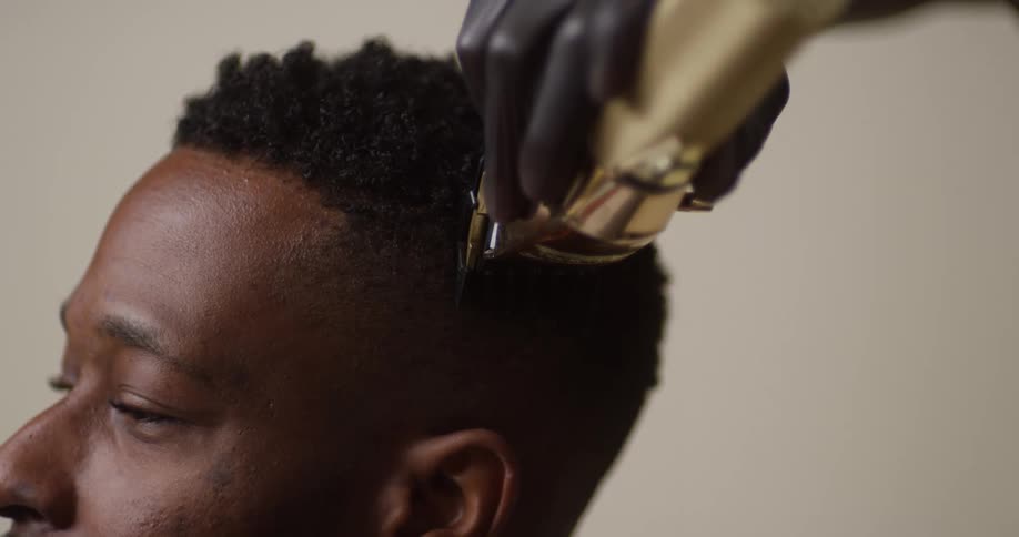 A hair stylist  wearing gloves trims a young, black man's hair with electric razor.