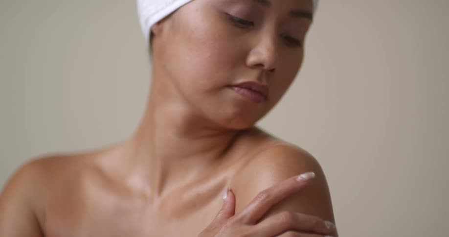 An Asian woman fresh out of the shower and with a towel on her head rubs lotion on her shoulder
