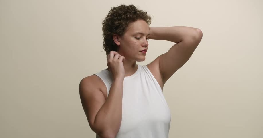 A young, white woman with short, curly hair modeling for the camera with one hand up in her hair and the other below her ear.