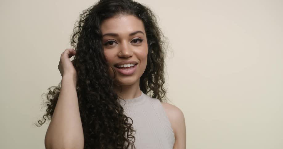 A young, exotic woman pulls her curly, dark hair over her shoulder to the front and gives a coy smile to the camera while adusting poses.