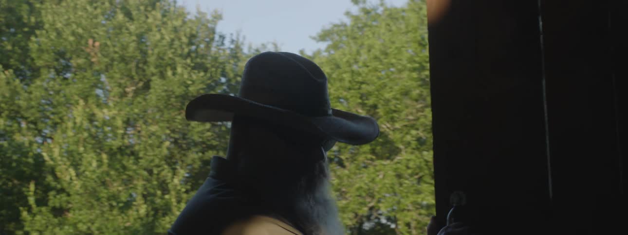 A man with a hat and sunglasses opening up a barn with trees the sky and clouds in the background