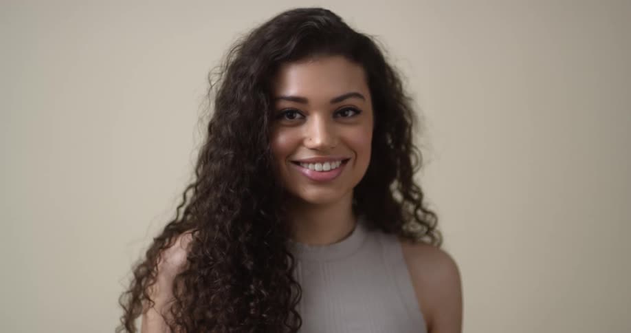 A young, ethnic female model pushes her dark, curly hair back, gives a coy smile to the camera, and then slowly looks off into the distance and back again.