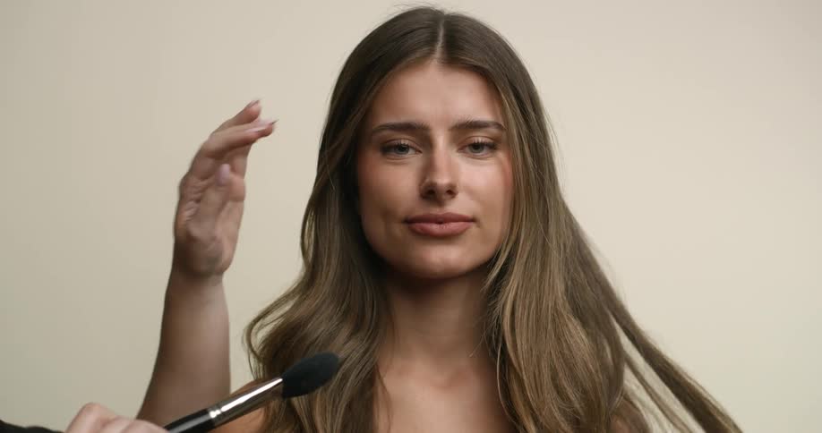 A young, white female model purses her lips in a tight, pinched smile as she is made up by stylists for a photo shoot